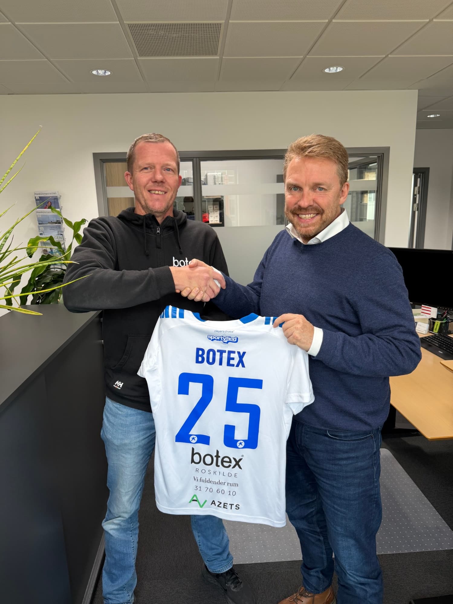 Sponsor og klubrepræsentant giver hånd og holder en spillertrøje med sponsorlogo.